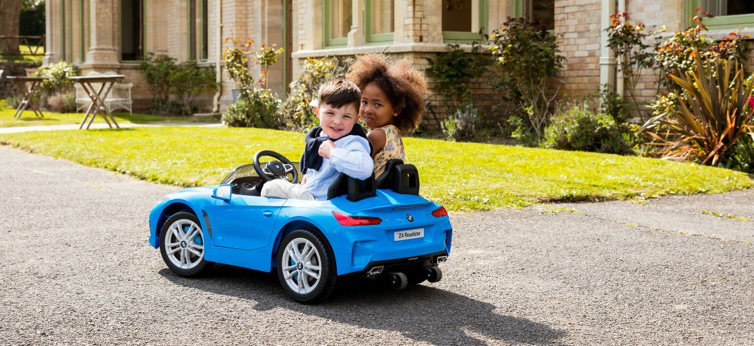 Two young children outside a large country house sat in a blue BMW ride on car