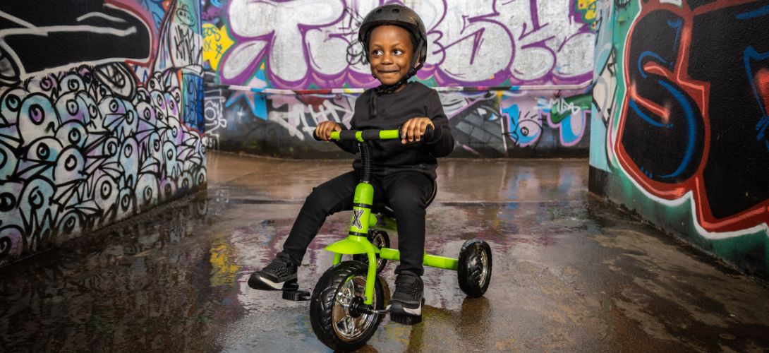 A young boy riding a green trike in through a graffiti'd tunnel