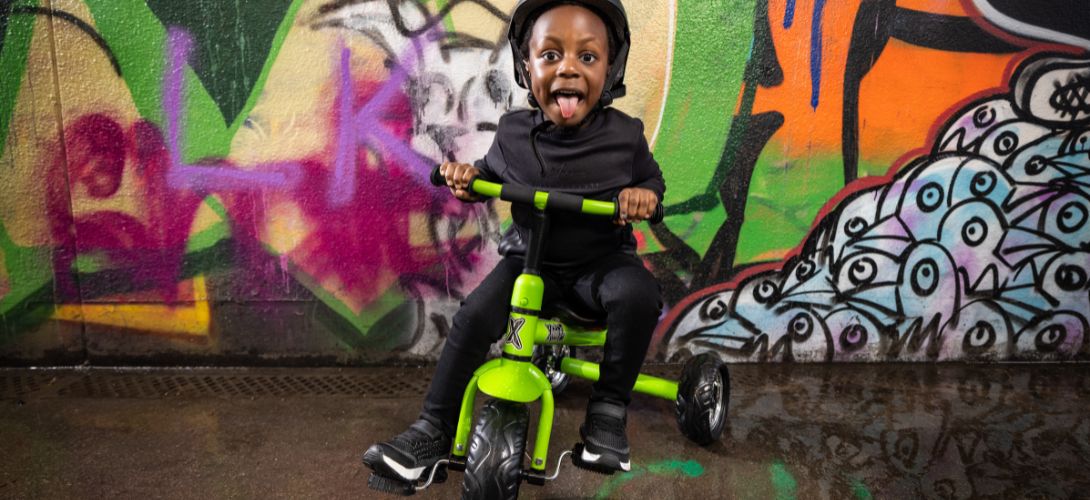 A young boy riding a green trike. He is cheekily sticking his tongue out. The backdrop is filled with orange and green graffiti.