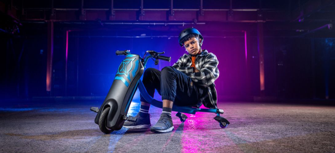 A boy sat on a blue and back Cyclone trike inside a warehouse lit with pink and blue lighting