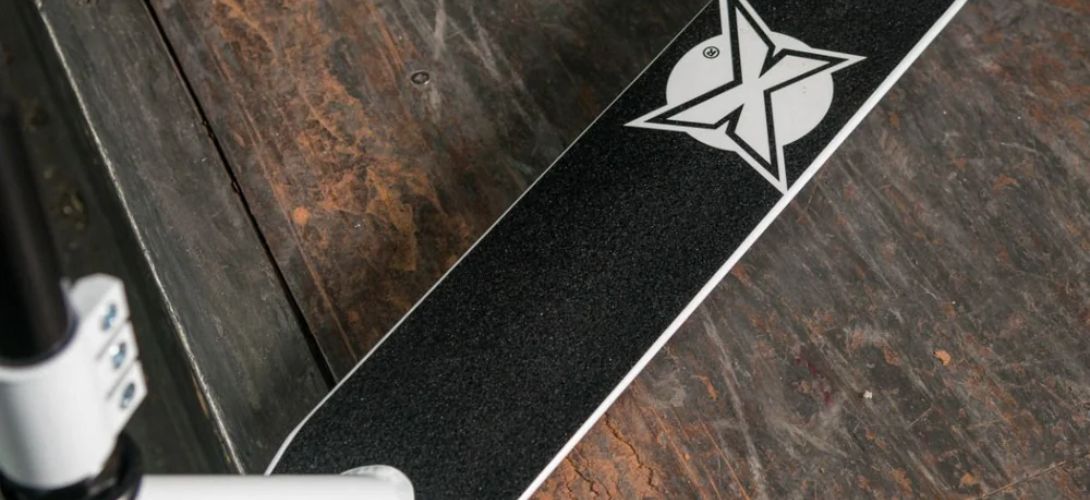 the deck of a white and black scooter against a wooden background. The black grip tape features a white Xootz X logo