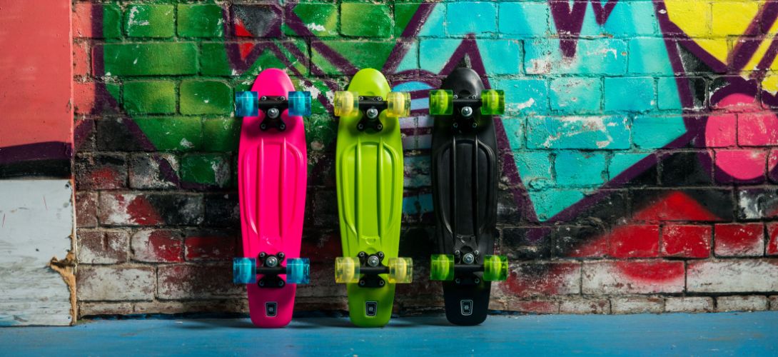 Three coloured cruiser skateboards - neon pink, green and black - stood upright against a graffiti'd brick wall